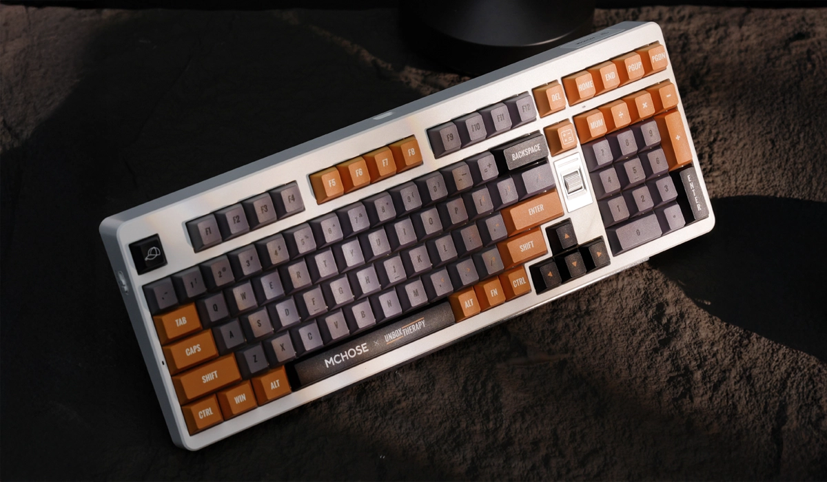 Klawiatura MCHOSE UT98 z szarymi i pomarańczowymi klawiszami leży na ciemnym tle.